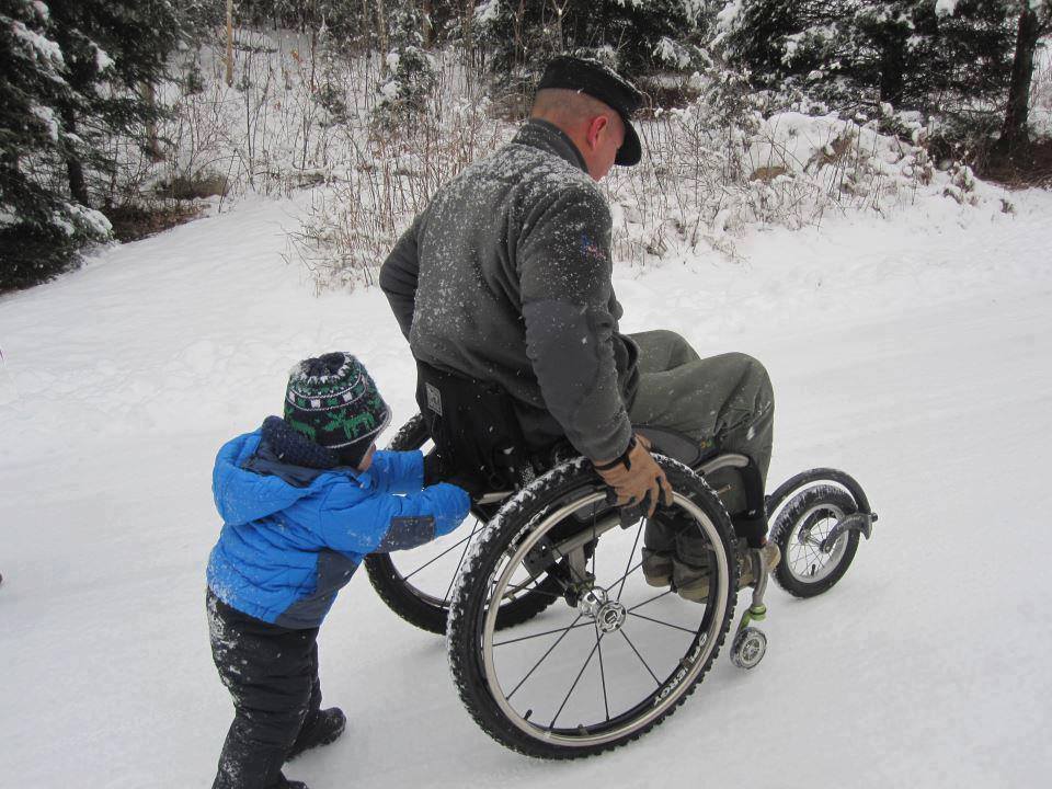 Un petit garcon pousse le fauteuil roulant de son papa dans la neige